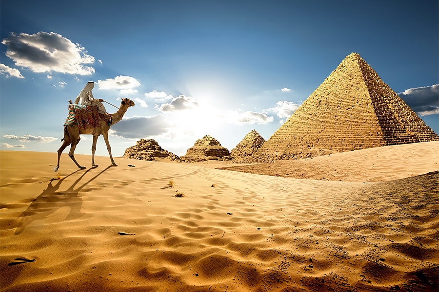 Pustynia. Po lewej stronie wielbłąd. Na wielbłądzie siedzi osoba ubrana w białe szaty. Po prawej stronie piramidy.