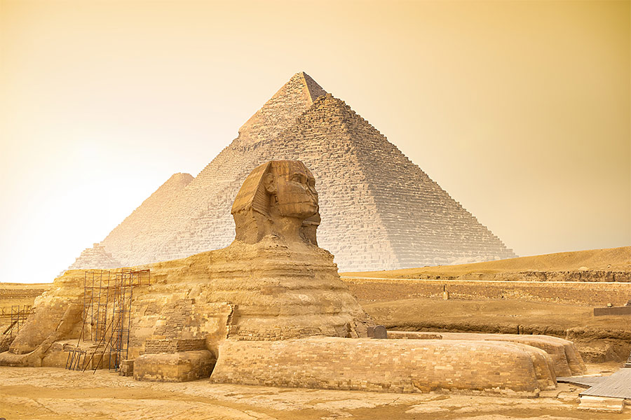 W centralnej części zdjęcie znajduje się Sfinks. Za nim piramidy. Niebo jest przysłonięte przez piasek. Ma kolor żółty.