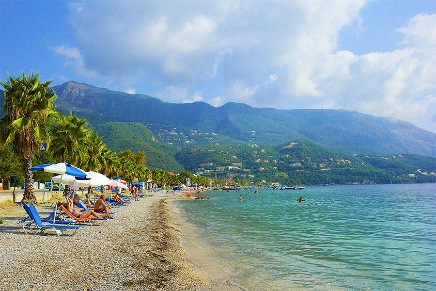 Po prawej plaża z leżakami, parasolami i palmami. Po prawej ludzie kąpią się w morzu. W tle góry.