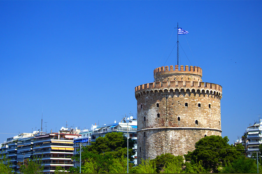 Po prawej wieża z flagą Grecji. Po lewej budynki. Niebo jest błękitne.